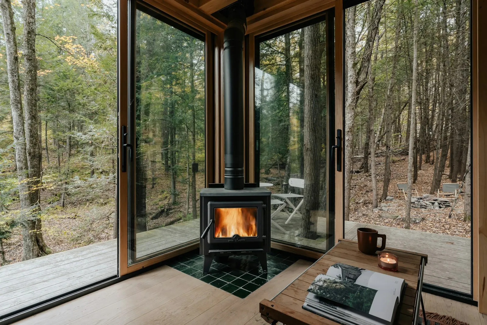 A lit wood stove in a cabin corner, floor-to-ceiling windows onto autumn forest