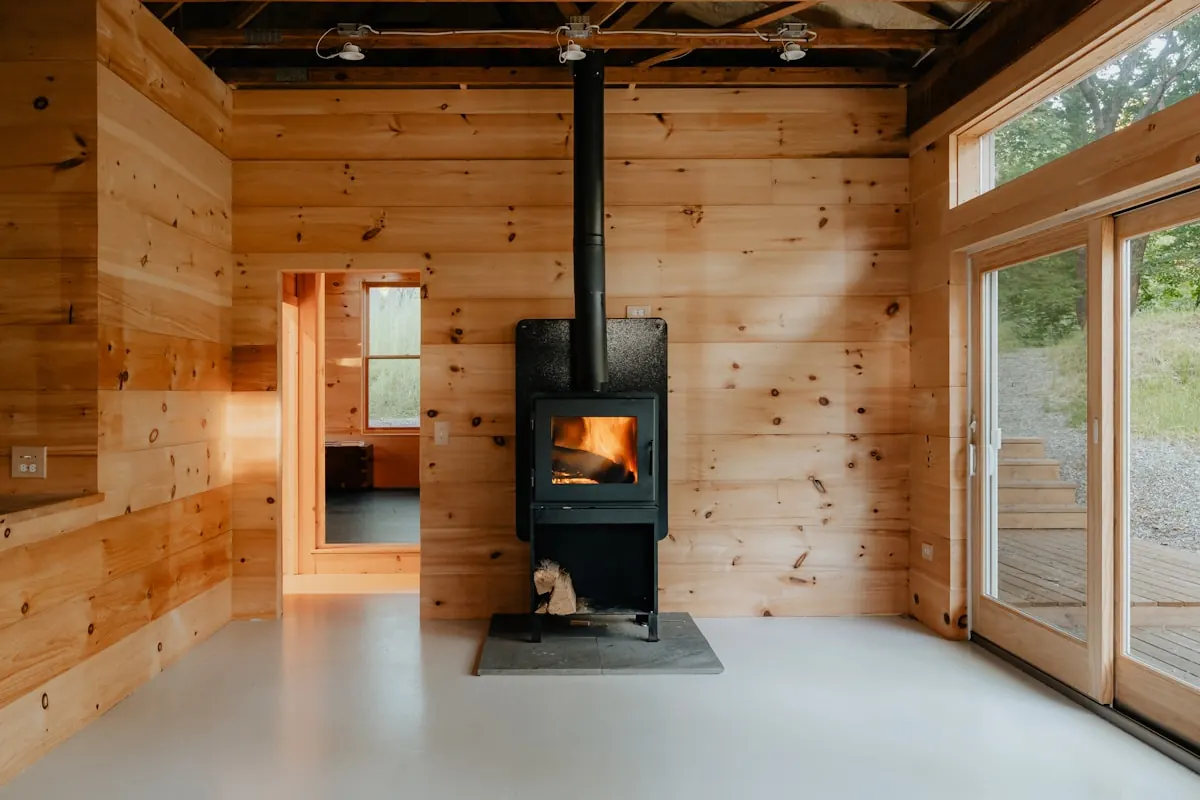 Pale timber room with a black wood stove and a wall of glass to the trees
