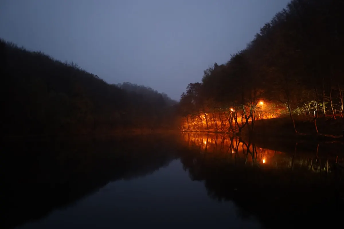 Still lake at twilight with dark forested hills and a small light on the shore